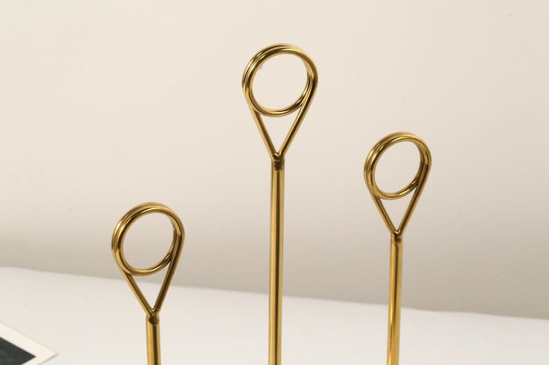 May include: Three gold-colored metal place card holders. Each holder has a slender stem that leads to a triangular shape, topped with a circular ring. The holders are arranged in a row, with varying heights.