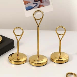 May include: Gold-colored metal place card holders with a circular base and a loop at the top. The holders are designed to display photos or cards. The image shows several holders, some with cards inserted, on a white surface.