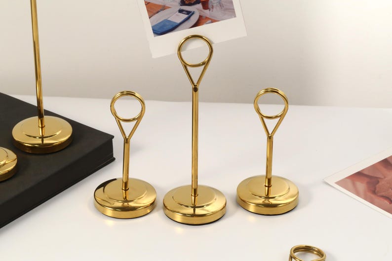 May include: Gold-colored metal place card holders with a circular base and a loop at the top. The holders are designed to display photos or cards. The image shows several holders, some with cards inserted, on a white surface.