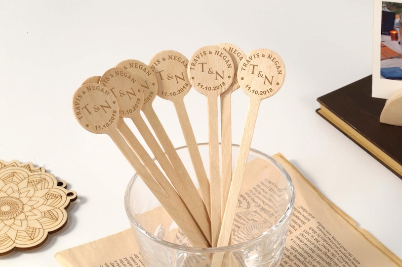 Puede incluir: Un conjunto de palitos de madera para remover bebidas con texto grabado. Cada palito tiene una parte superior circular con los nombres "Travis & Negan", las iniciales "T&N" y la fecha "11.10.2018".