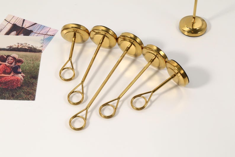 May include: A collection of gold-colored metal photo holders. Each holder has a circular base, a slender stem, and a circular top. The holders are arranged on a white surface, with a photo in the background.