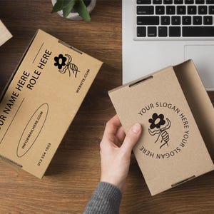 May include: Two open brown cardboard boxes with custom printed text and a floral design. One box has the text "YOUR NAME HERE ROLE HERE" and contact information. The other box has the text "YOUR SLOGAN HERE". The boxes are on a wooden surface.