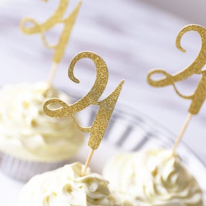 以下が含まれることがあります： ゴールドのグリッターで「21」と書かれたケーキトッパーがついたカップケーキが3つ。カップケーキは白いフロスティングで覆われています。