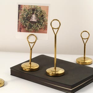 May include: Four gold-colored metal photo holders with a circular top and a round base. One holder displays a photo of a wreath. The holders are arranged on a black book and a white surface.