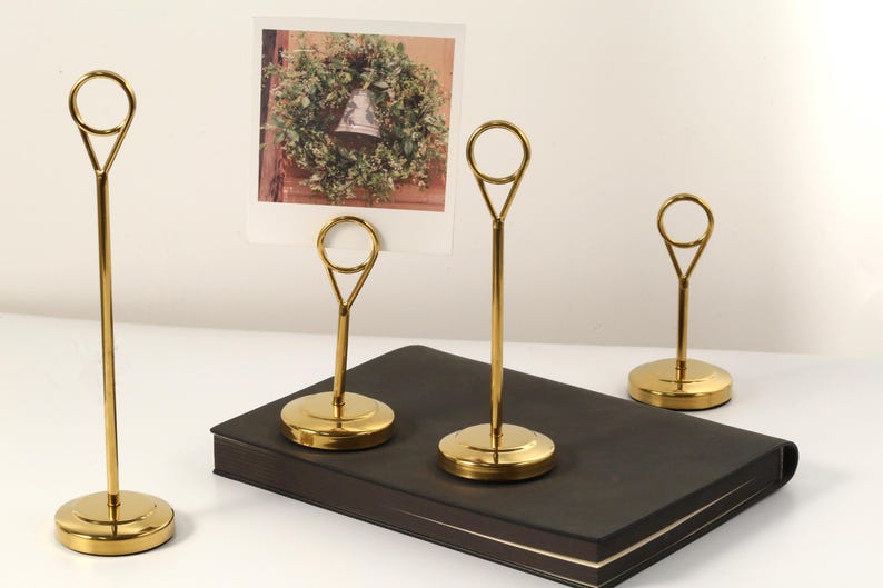 May include: Four gold-colored metal photo holders with a circular top and a round base. One holder displays a photo of a wreath. The holders are arranged on a black book and a white surface.