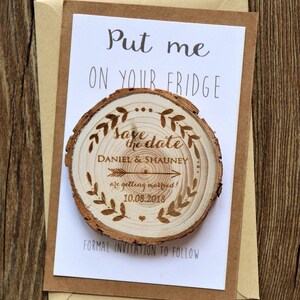 May include: A wooden save the date magnet with a rustic woodgrain finish. The magnet features a wood-burned design with the text "Save the date" and the names "Daniel & Shauney" with an arrow pointing to the date "10.08.2018". The magnet is attached to a white card with the text "Put me on your fridge" and "Formal invitation to follow".