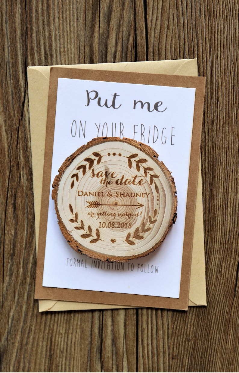 May include: A wooden save the date magnet with a rustic woodgrain finish. The magnet features a wood-burned design with the text "Save the date" and the names "Daniel & Shauney" with an arrow pointing to the date "10.08.2018". The magnet is attached to a white card with the text "Put me on your fridge" and "Formal invitation to follow".