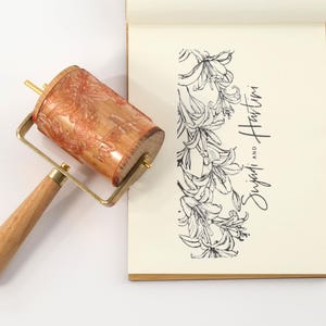May include: A wooden print roller with a patterned design, brass hardware, and a wooden handle. Next to it is a notepad with a floral illustration and the text "Sujadi and Hartini" in black script.