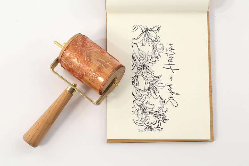 May include: A wooden print roller with a patterned design, brass hardware, and a wooden handle. Next to it is a notepad with a floral illustration and the text "Sujadi and Hartini" in black script.