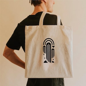 May include: A beige canvas tote bag featuring a black graphic design of a cactus with a rainbow, heart, and stars. The bag has two long straps and is carried over a person's shoulder.