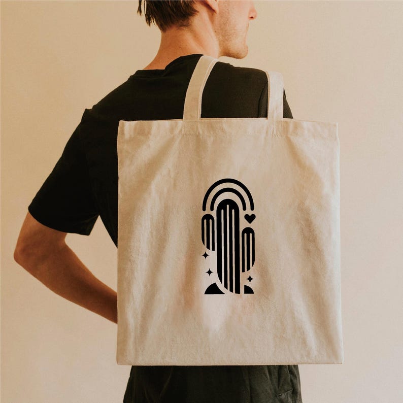 May include: A beige canvas tote bag featuring a black graphic design of a cactus with a rainbow, heart, and stars. The bag has two long straps and is carried over a person's shoulder.