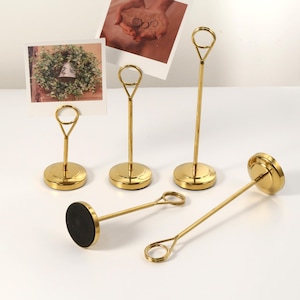 May include: A collection of gold-colored metal photo holders. The holders have a circular base, a slender stem, and a loop at the top for holding photos or cards. Some holders are upright, while others are lying down.