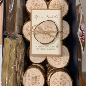 May include: A close-up of a box filled with wooden save-the-date magnets. The magnets are round and feature a rustic wood grain design. Each magnet has the text "Save the Date" and the couple's names and wedding date. The text "You're Invited!" is printed on a card in the center of the image.