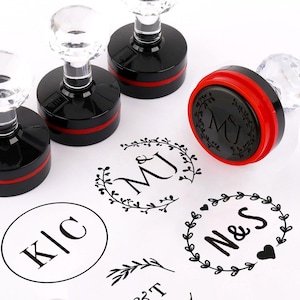 Puede incluir: Sellos de goma negros y rojos con mangos de cristal transparente. Los sellos están junto a varios diseños estampados en tinta negra, incluyendo iniciales dentro de coronas y una ramita simple. Los sellos son para crear diseños personalizados.