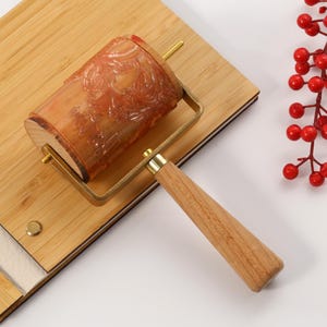May include: A wooden roller with a carved design, brass hardware, and a wooden handle rests on a bamboo cutting board. A sprig of red berries is to the right. The roller is likely used for crafts or baking.
