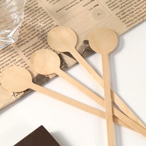 Puede incluir: Una colecci&oacute;n de peque&ntilde;as cucharas de madera de color claro con cabezas redondas y mangos largos y delgados. Las cucharas est&aacute;n dispuestas sobre un fondo de peri&oacute;dico, lo que sugiere un tema artesanal o culinario.