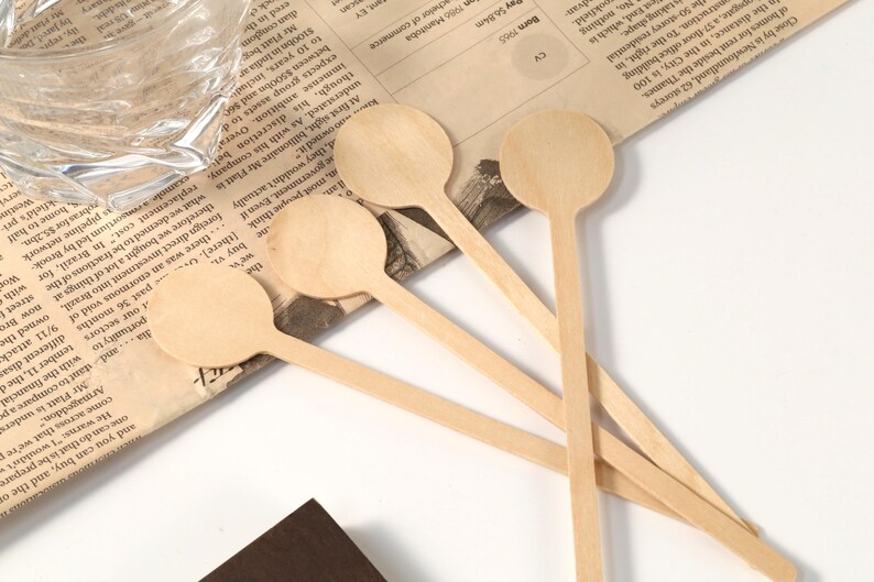 Puede incluir: Una colecci&oacute;n de peque&ntilde;as cucharas de madera de color claro con cabezas redondas y mangos largos y delgados. Las cucharas est&aacute;n dispuestas sobre un fondo de peri&oacute;dico, lo que sugiere un tema artesanal o culinario.
