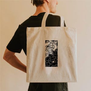 May include: A beige tote bag with a black and white floral design. The rectangular graphic features detailed line art of roses and leaves. The bag has two long straps and is carried over the shoulder.