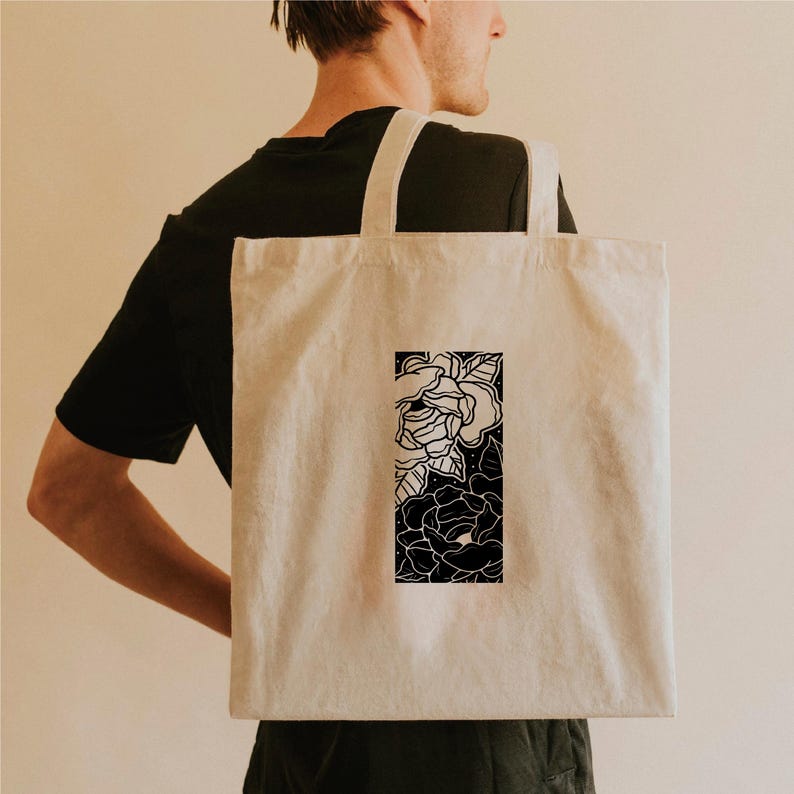 May include: A beige tote bag with a black and white floral design. The rectangular graphic features detailed line art of roses and leaves. The bag has two long straps and is carried over the shoulder.