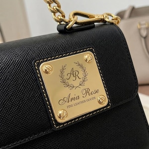 以下が含まれることがあります： 黒いテクスチャードレザーのハンドバッグ。Aria Rose FINE LEATHER GOODSの文字と月桂樹の冠に囲まれたARのモノグラムが刻印されたゴールドトーンの金属プレートが付いています。バッグにはロブスタークラスプで取り付けられたゴールドのチェーンストラップが付いています。
