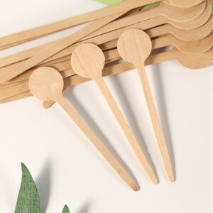 Puede incluir: Un conjunto de peque&ntilde;as cucharas de madera de color claro, con cabezas redondas y mangos largos y finos. Varias cucharas est&aacute;n apiladas, mientras que otras est&aacute;n dispuestas sobre una superficie blanca, con hojas verdes en primer plano.