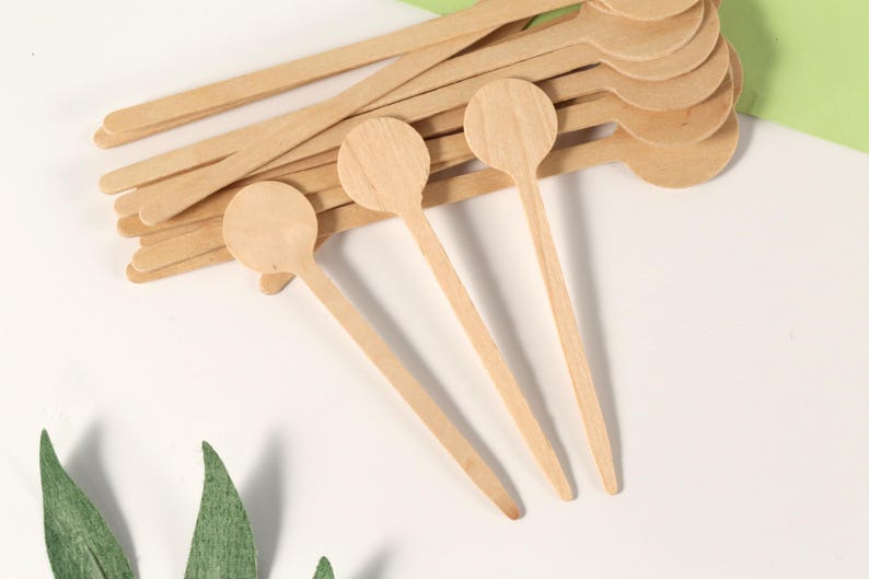 Puede incluir: Un conjunto de peque&ntilde;as cucharas de madera de color claro, con cabezas redondas y mangos largos y finos. Varias cucharas est&aacute;n apiladas, mientras que otras est&aacute;n dispuestas sobre una superficie blanca, con hojas verdes en primer plano.
