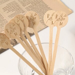 Puede incluir: Un conjunto de palitos de madera para remover con dise&ntilde;os florales grabados y las letras "R" y "M". Los palitos est&aacute;n dispuestos en un cuenco de cristal transparente, con un peri&oacute;dico vintage y un libro encuadernado en cuero en el fondo. Los palitos son de madera clara.