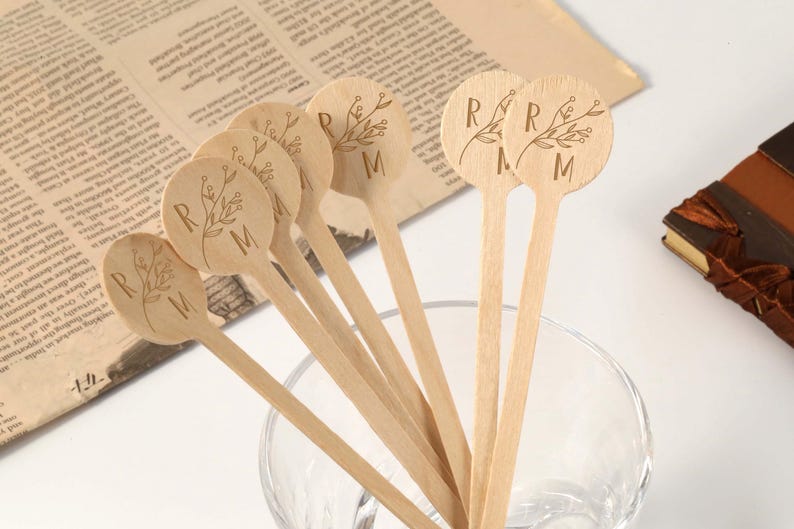 Puede incluir: Un conjunto de palitos de madera para remover con dise&ntilde;os florales grabados y las letras "R" y "M". Los palitos est&aacute;n dispuestos en un cuenco de cristal transparente, con un peri&oacute;dico vintage y un libro encuadernado en cuero en el fondo. Los palitos son de madera clara.