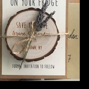 May include: A rustic save the date announcement with a wooden slice tied with twine and a sprig of lavender. The text reads "Save the Date" with the names Lerane and Brandon, the date 19, and the location "TAMOWN, NY". The announcement also includes the text "Formal Invitation to Follow".