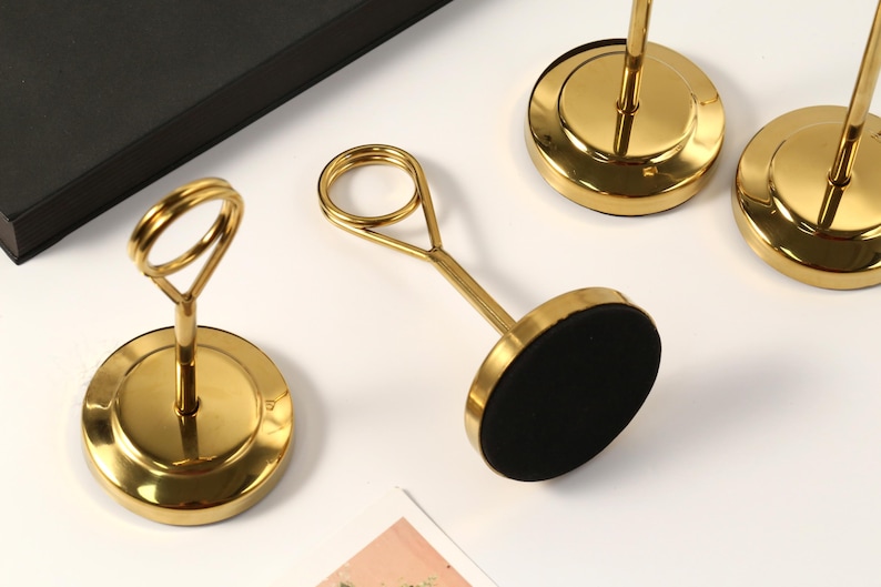 May include: Gold-colored metal place card holders with a circular base and a loop at the top. The base has a black, circular pad. The holders are arranged on a white surface, with a black box in the background.