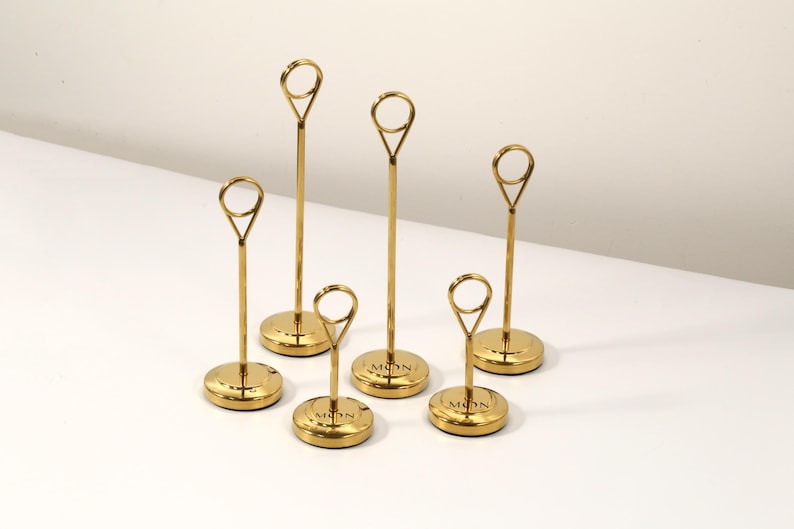 May include: Six gold-colored place card holders. Each holder has a circular base with the word "MON" engraved on it, a slender stem, and a loop at the top for holding a card. The holders are arranged on a white surface.