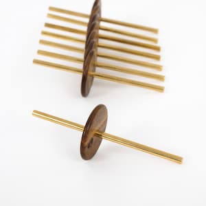 Peut inclure: Un ensemble de sept fuseaux, chacun composé d'une fusaïole circulaire en bois brun foncé centrée sur une longue tige fine en laiton poli. Un fuseau est placé au premier plan, tandis que les six autres sont empilés en rangée parallèle derrière.