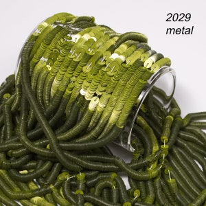May include: Olive green metal sequins spilling from a clear glass container. The sequins are round and strung together, creating a textured, flowing effect. The text "2029 metal" is visible in the upper right corner.