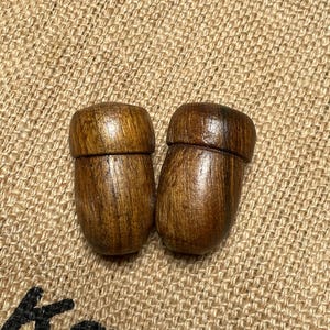 May include: Two acorn-shaped wooden objects with a natural wood grain finish. The acorns are a medium brown colour and have a smooth, polished appearance. They are set against a textured, tan burlap background.