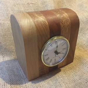 May include: A wooden clock with a gold-coloured face and Roman numeral markings. The clock is made of different types of wood, creating a striped pattern.