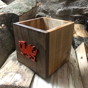 May include: A two-tone wooden box featuring a red dragon emblem. The box is square-shaped, with a darker wood front and lighter wood sides. The dragon emblem is a detailed, stylized representation of the Welsh dragon.