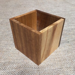May include: A square wooden box with a light brown wood exterior and a darker brown wood interior. The box is made of two different types of wood.