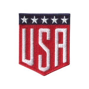 May include: A shield-shaped embroidered patch featuring a dark blue top band with five white stars. The lower section is red with the white embroidered letters USA centered in a bold, block font. The patch has a pointed bottom edge.