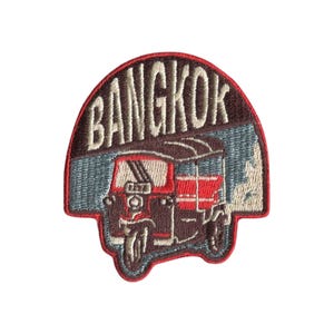 May include: An embroidered patch featuring a brown and red tuk-tuk vehicle against a blue background. The word BANGKOK is stitched in large, light-colored capital letters across the top of the patch, which is outlined in a bold red border.