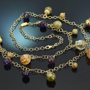Könnte beinhalten: Eine goldene Kettenhalskette mit einer Reihe hängender Anhänger, darunter facettierte violette Amethystperlen, durchscheinende gelbe Citrin-Kugeln und blassgrüne Edelstein-Orbs, durchsetzt mit kleinen, texturierten goldenen Filigran-Metallkugeln.