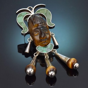 Könnte beinhalten: Vintage Anhänger mit einem geschnitzten Gesicht mit Kopfschmuck, akzentuiert mit grünen und silbernen Details. Der Anhänger hat hängende Elemente mit Tigeraugenperlen und silbernen Kappen.