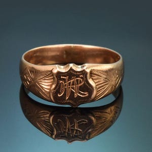 May include: A rose gold ring featuring a central shield-shaped plaque engraved with the intertwined initials JR. The band is decorated with etched fan-like patterns on both sides of the central shield, creating a detailed and textured appearance.