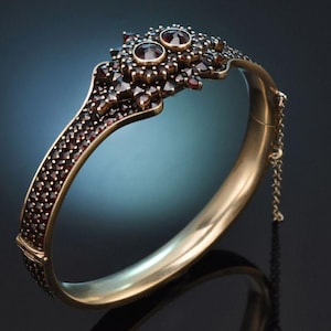 Peut inclure: Un bracelet doré à charnière avec un design détaillé. Le bracelet est orné de nombreux petits gemmes rouge foncé et de deux gemmes plus grandes en haut. Une fine chaîne sécurise le fermoir.
