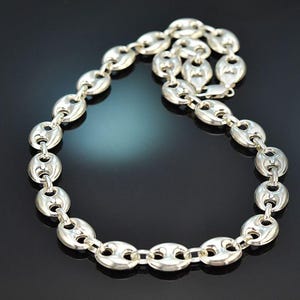 Könnte beinhalten: Eine silberfarbene Halskette bestehend aus einer Reihe polierter, ovaler, gewölbter Glieder, die durch kleinere kreisförmige Ringe verbunden sind. Die Kette hat eine reflektierende, metallische Oberfläche und ist in einer Schlaufe angeordnet.