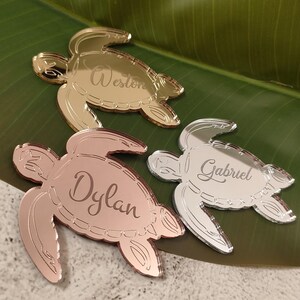 May include: Three mirrored turtle-shaped place cards. One is gold, one is rose gold, and one is silver. Each turtle has a name engraved on its shell: Weston, Dylan, and Gabriel. The place cards are on a green leaf.