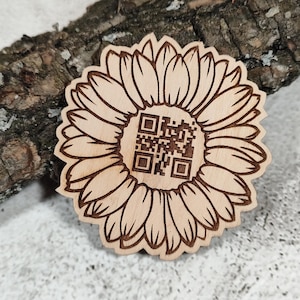 May include: A light-colored wooden sunflower cutout featuring a dark brown laser-engraved QR code in the center. The sunflower has numerous detailed petals radiating from the middle, with the square QR code clearly visible on the flat surface.