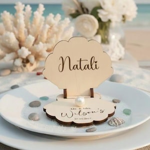 Peut inclure: Un porte-nom en bois en forme de coquillage avec le nom "Natali" écrit en script. Il est posé sur une assiette blanche avec du sable, des galets et une perle. La base indique "Mr. & Mrs. Wilson's 07.05.2027".