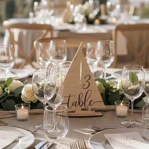 May include: A beautifully set table for a wedding or event. The centerpiece is a wooden table number with the number 3 and the word "table" in elegant script. White roses and greenery adorn the table, along with glassware, candles, and place settings.