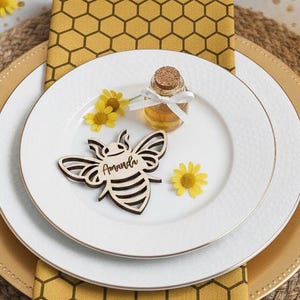 Könnte beinhalten: Ein Tischarrangement zum Thema Biene. Es umfasst einen weißen Teller mit einer bienenförmigen Namenskarte, eine kleine Honigflasche und eine gelbe Bienenwaben-Serviette. Ein Kuchenstück und ein gelbes Glas sind ebenfalls zu sehen.