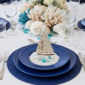 May include: A dining table setting featuring dark blue plates, clear glassware, and silver cutlery. Each place setting includes a small wooden sailboat cutout with the name "Raven" written on it, resting on a bed of white sand and blue glass beads.